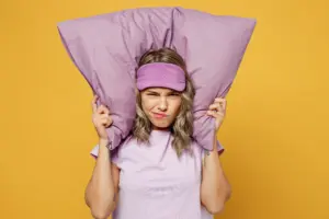 Sad tired young woman she wearing purple pyjamas jam sleep eye mask at home covering ears from neighbours noise snoring isolated on plain yellow background studio portrait. Bad mood night nap concept